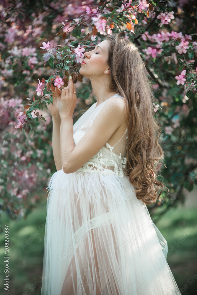 Pregnant pretty woman is wearing white fashion dress posing in pink blossom apple garden, summer time Pregnant pretty woman is wearing white fashion dress posing in pink blossom apple garden, summer time