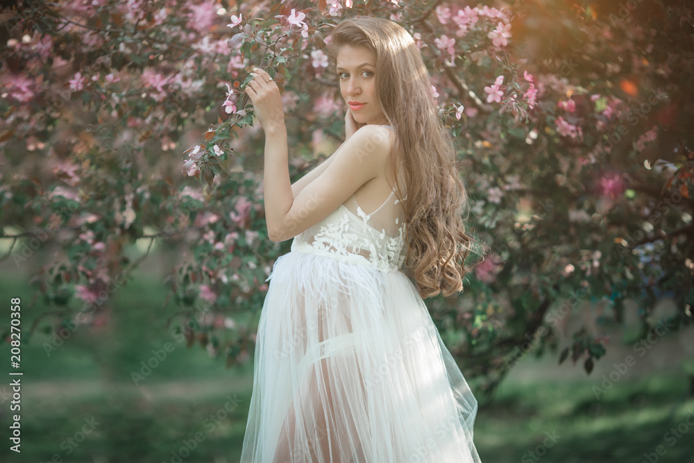 Pregnant pretty woman is wearing white fashion dress posing in pink blossom apple garden, summer time Pregnant pretty woman is wearing white fashion dress posing in pink blossom apple garden, summer time
