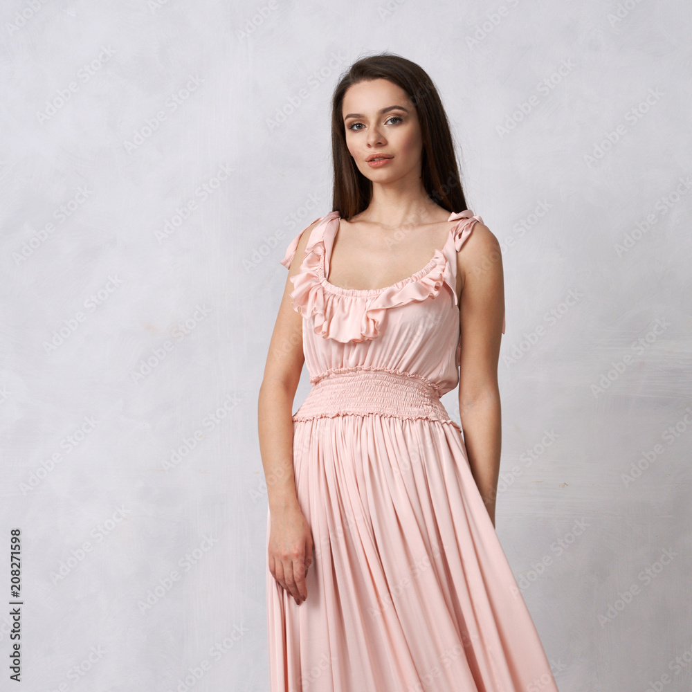 Full body portrait of gorgeous young brunette woman dressed in exquisite nude ball gown with lace top. Attractive female model in elegant strapless dress posing against white wall on background.