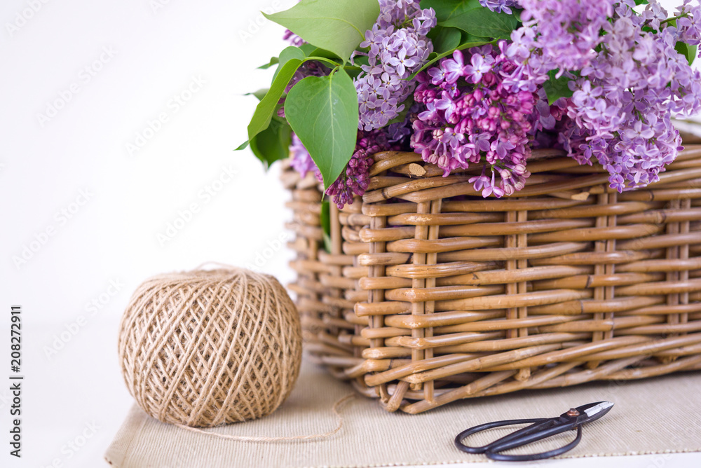 Lilac spring bouquet in wooden basket on table with scissors and rope on white wall background