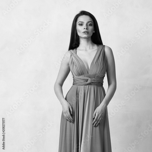 Beautiful long haired young woman dressed in stylish red bandeau maxi dress posing against white wall on background. Elegant brunette female model demonstrating evening outfit in studio.