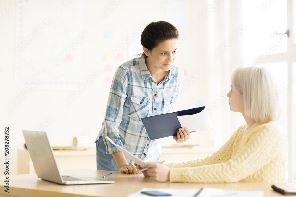 Young blonde female sitting at desk with laptop holding papers talking to brunette standing near and smiling Young blonde female sitting at desk with laptop holding papers talking to brunette standing near and smiling