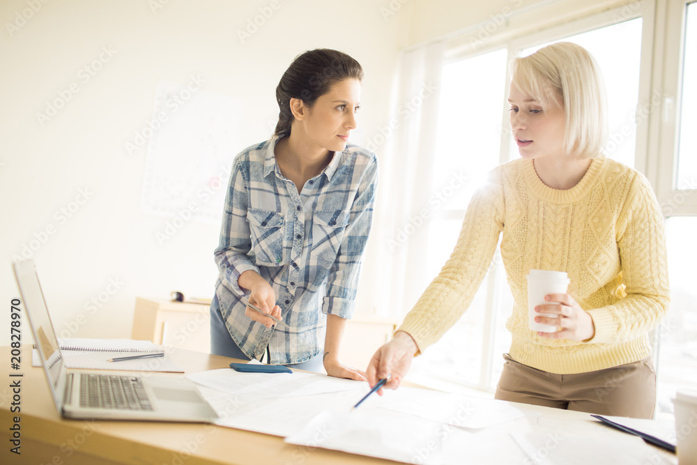 Young female office workers talking standing at desk pointing at papers Young female office workers talking standing at desk pointing at papers