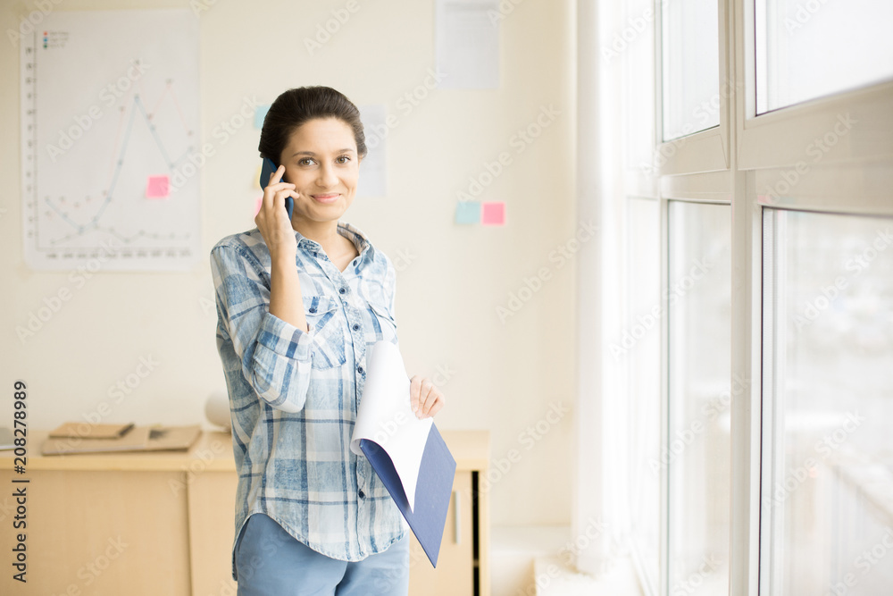 Young pretty female standing near window at office talking on phone holding folder smiling and looking at camera Young pretty female standing near window at office talking on phone holding folder smiling and looking at camera