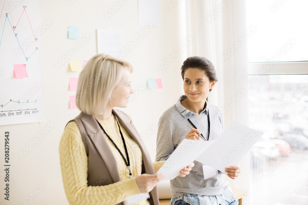 Side view of pretty smiling ladies with badge on neck holding papers in hands and looking at each other Side view of pretty smiling ladies with badge on neck holding papers in hands and looking at each other