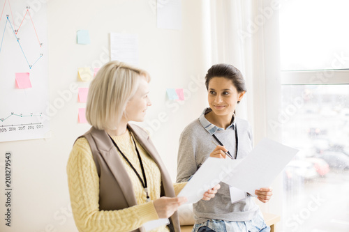 Side view of pretty smiling ladies with badge on neck holding papers in hands and looking at each other