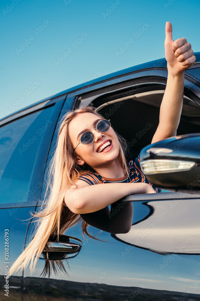 Naklejka premium smiling stylish woman in sunglasses leaning out from car window and doing thumb up gesture