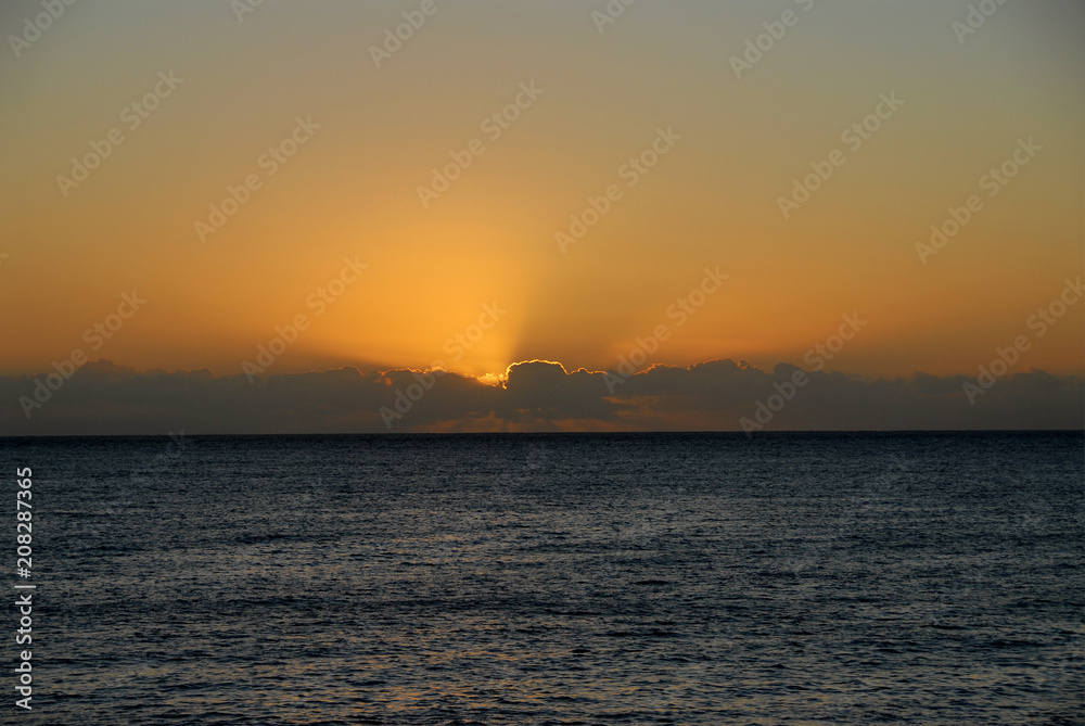 ssun setting into dark clouds against orange sky above ocean