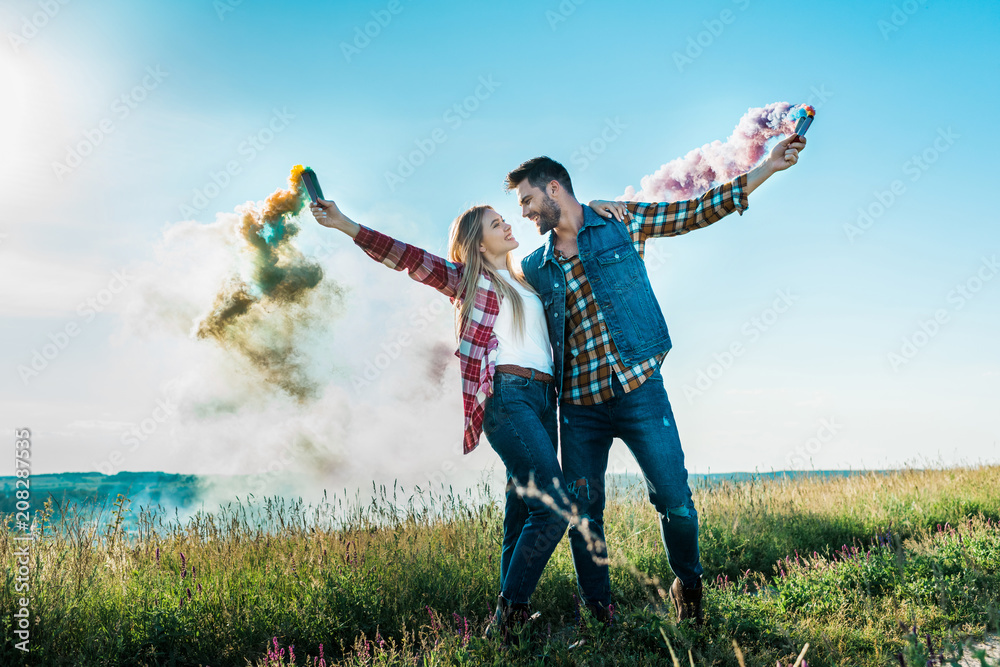 young couple looking at each other and holding colorful smoke bombs on rural meadow young couple looking at each other and holding colorful smoke bombs on rural meadow