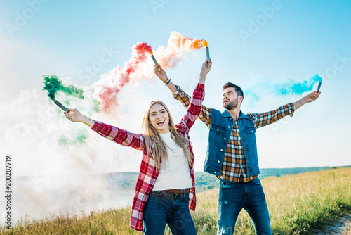 smiling young couple holding colorful smoke bombs on rural meadow