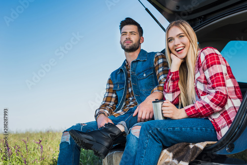 smiling woman with coffee cup and her boyfriend sitting near on car trunk in field © LIGHTFIELD STUDIOS