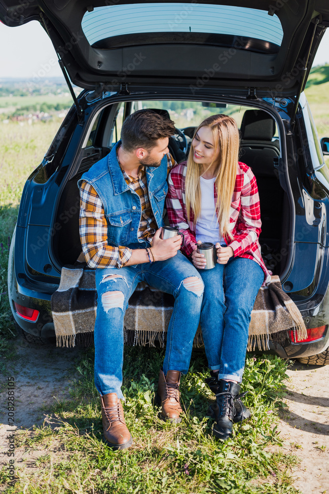 couple of stylish tourists with coffee cups sitting on car trunk in rural field