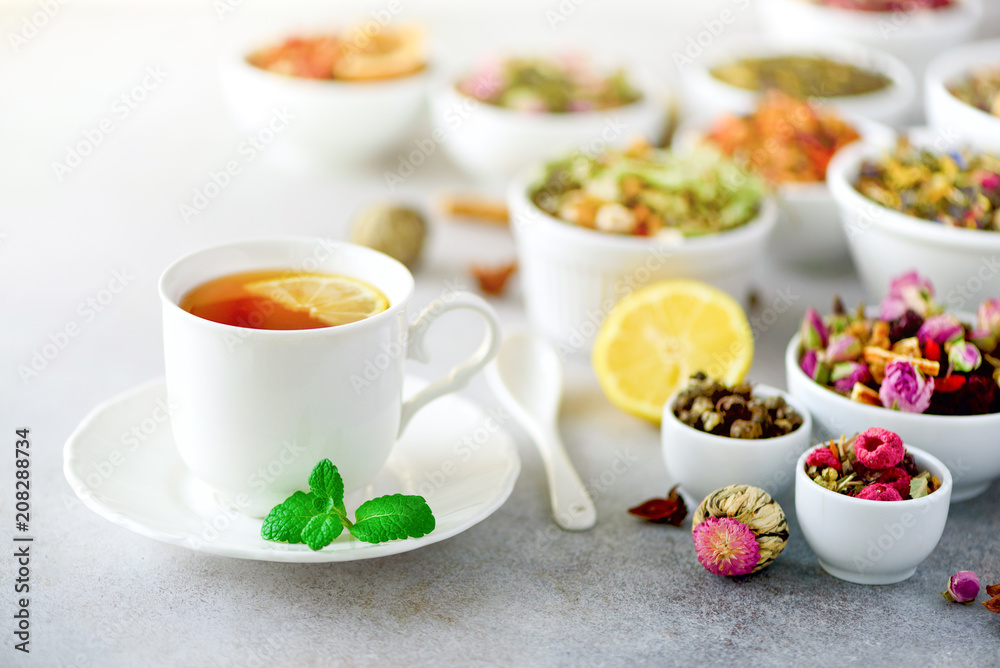 Tea concept with copy space. Different kinds of dry tea in white ceramic bowls and cup of aromatic tea on grey background. Tea concept with copy space. Different kinds of dry tea in white ceramic bowls and cup of aromatic tea on grey background.