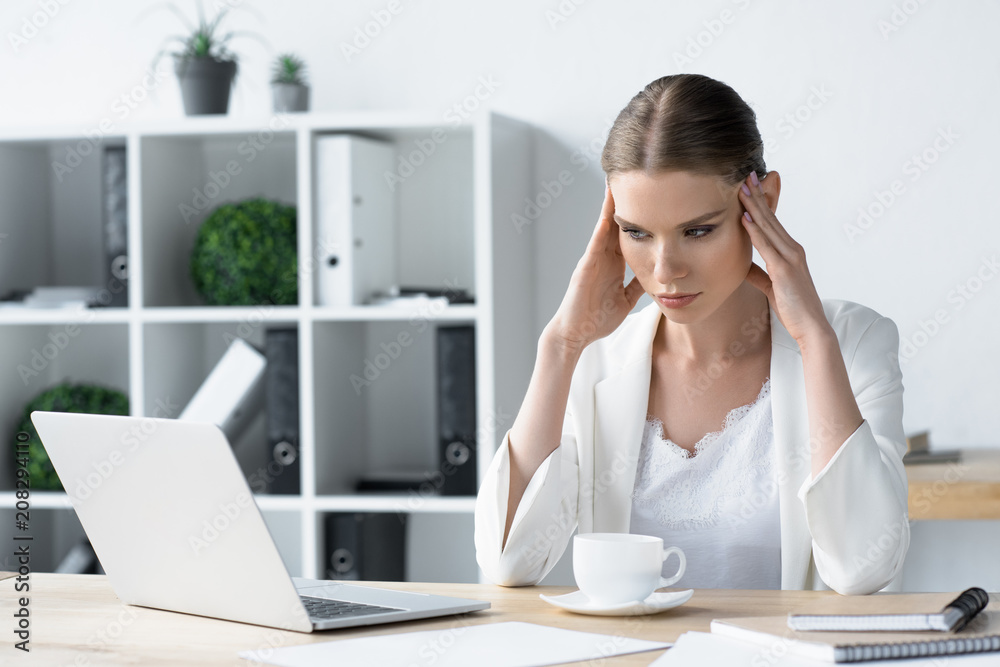overworked young businesswoman with headache sitting at workplace in office and holding head