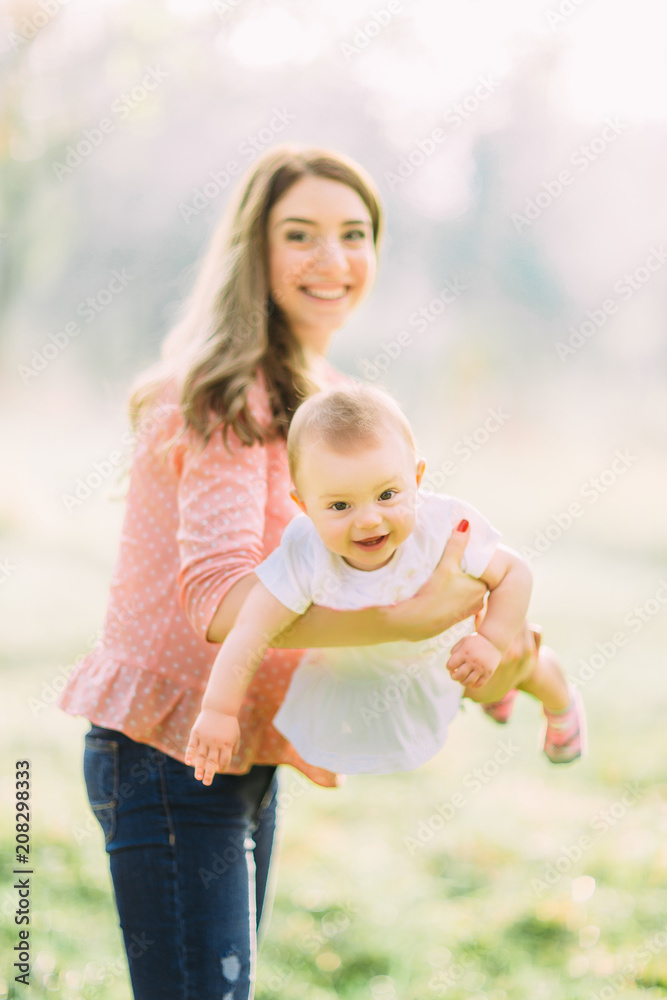 Fototapeta premium Young mother holding daughter in her arms. Beautiful Mother And Baby outdoors. Nature. Beauty Mum and her Child playing in Park together. Outdoor Portrait of happy family. Joy. Mom and Baby