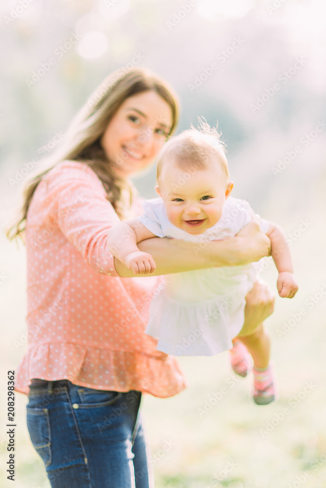 Fototapeta premium Young mother holding daughter in her arms. Beautiful Mother And Baby outdoors. Nature. Beauty Mum and her Child playing in Park together. Outdoor Portrait of happy family. Joy. Mom and Baby