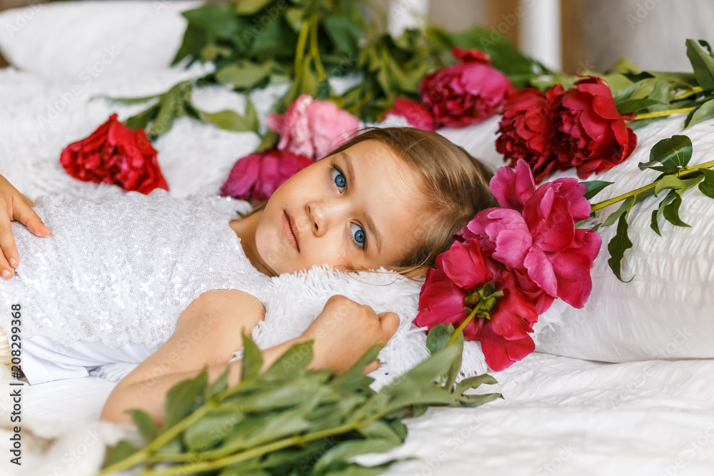 Cute little girl with peonies in studio lying on the bed among peonies Cute little girl with peonies in studio lying on the bed among peonies
