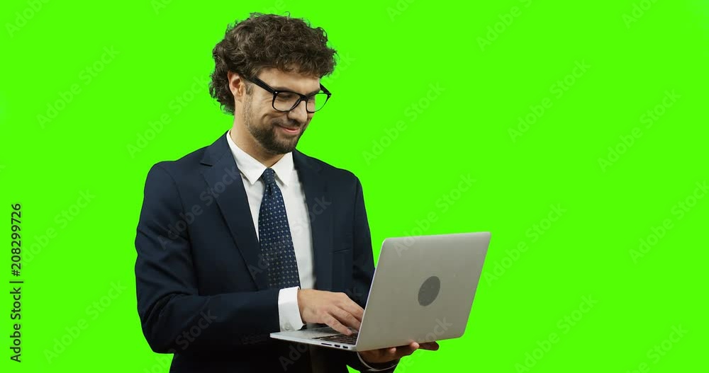 Attractive businessman in glasses and tie giving his thumb up while standing on the green screen background and working on the laptop. Chroma key. Attractive businessman in glasses and tie giving his thumb up while standing on the green screen background and working on the laptop. Chroma key.