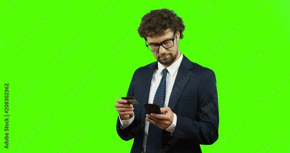 Attractive businessman in glasses and tie entering the credit card number on his smartphone while paying online and standing on the green screen background and working on the laptop. Chroma key. Attractive businessman in glasses and tie entering the credit card number on his smartphone while paying online and standing on the green screen background and working on the laptop. Chroma key.