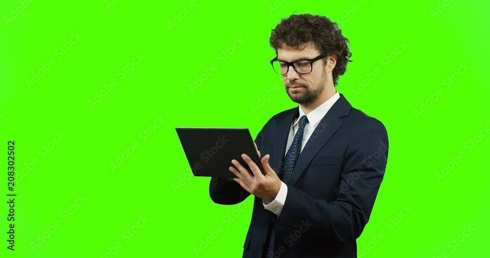 Caucasian man in glasses, suit and tie standing on the chroma key background and typing on the laptop, then smiling and posing to the camera. Green screen. Caucasian man in glasses, suit and tie standing on the chroma key background and typing on the laptop, then smiling and posing to the camera. Green screen.