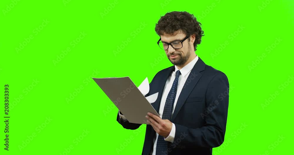Attractive smiled Caucasian businessman in the glasses, suit and tie looking at the documents in his hands and being satisfied on the green screen. Chroma key. Attractive smiled Caucasian businessman in the glasses, suit and tie looking at the documents in his hands and being satisfied on the green screen. Chroma key.