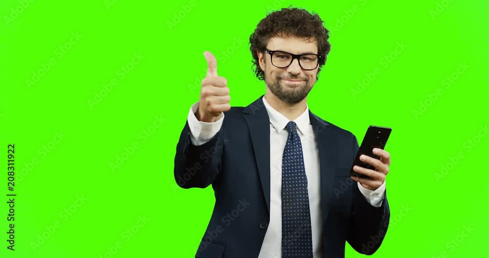 Caucasian smiled man in glasses, suit and tie giving his thumb up while scrolling and typing on the smartphone on the green screen background. Chroma key. Caucasian smiled man in glasses, suit and tie giving his thumb up while scrolling and typing on the smartphone on the green screen background. Chroma key.