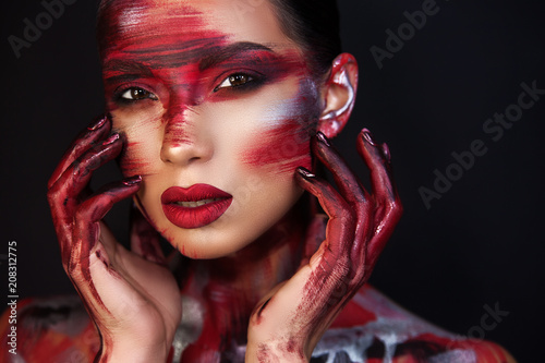 a beautiful girl with Asian roots on a black background holds her hands in the face which are covered with an original makeup with a red train.