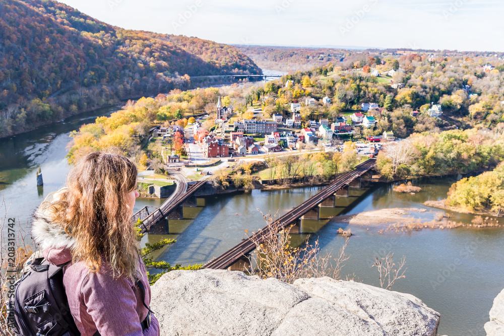 Hiker woman girl looking at cityscape overlook, colorful orange yellow foliage fall autumn forest with small village town by river in Harpers Ferry, West Virginia, WV