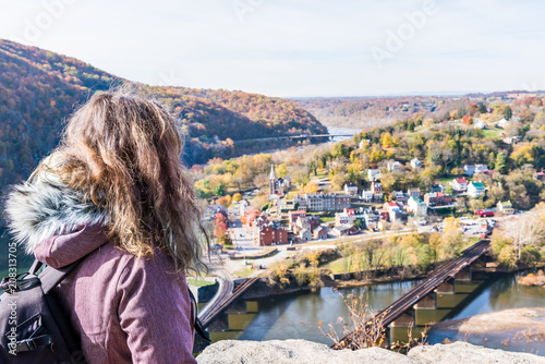 Overlook, hiker woman girl looking at cityscape, colorful orange yellow foliage fall autumn forest with small village town by river in Harpers Ferry, West Virginia, WV photo