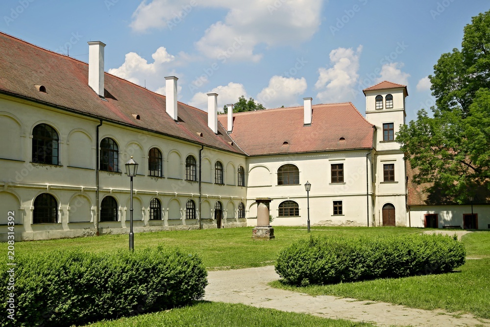 Fototapeta premium White building of former monastery with red roofs and chimneys standing in a green garden, sandy paths, lamps, today a museum in Predklasteri, close to Brno, Czech Republic, Europe