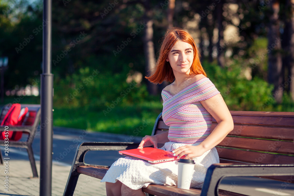 Naklejka premium A young red-haired woman in a white romantic skirt, a pink top and blue Sneakers sneakers holds a noubuk, looks around and sits on a bench on a summer day in the city park