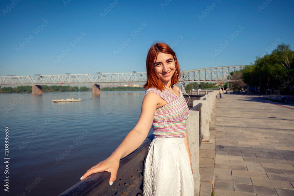 A young red-haired woman in a white romantic skirt, a pink top listens to music on the phone and looks at the river on the city waterfront on a summer day