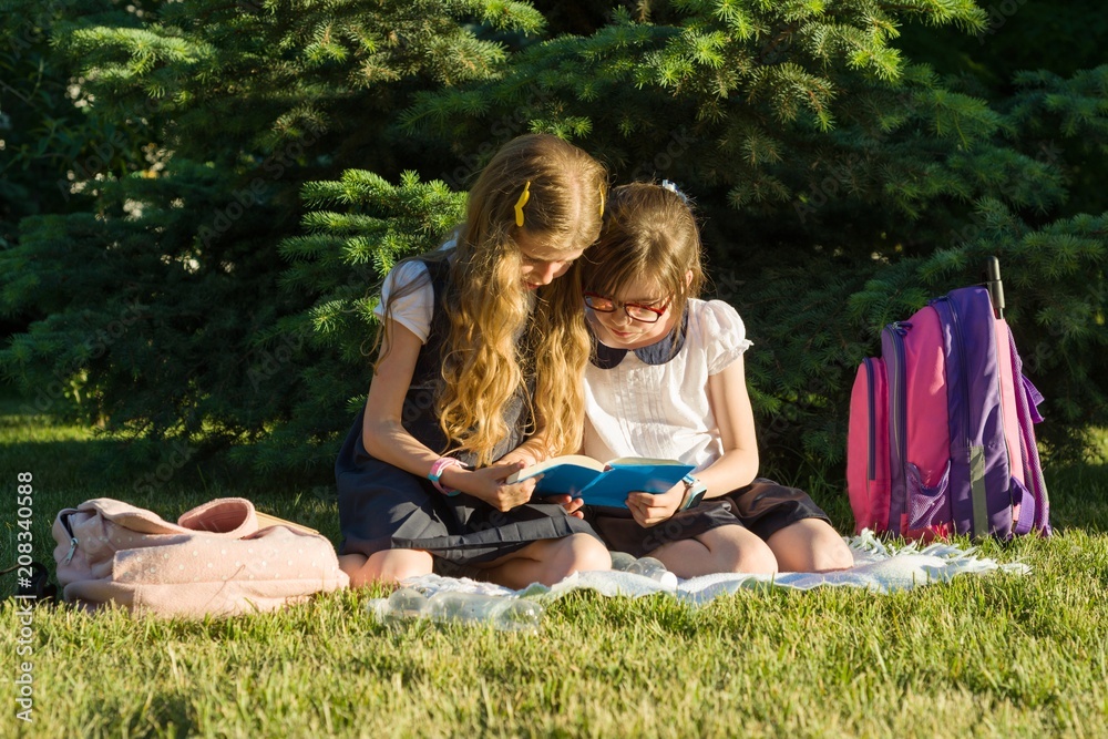 Two little girl friends schoolgirl learns sitting on a meadow in the park. Children with backpacks, books, notebooks. Two little girl friends schoolgirl learns sitting on a meadow in the park. Children with backpacks, books, notebooks.