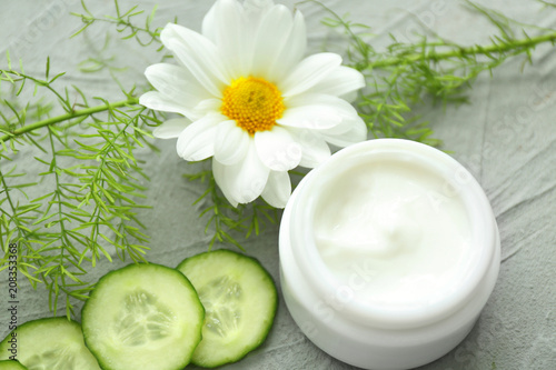 Jar of cream with herbal extract and cucumber slices on grey background