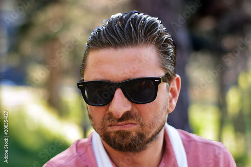 Young man's outdoors portrait looking at the camera who has sunglasses with natural green background. Neutral, happy, sad, disgust, fear, surprise Facial Emotion.