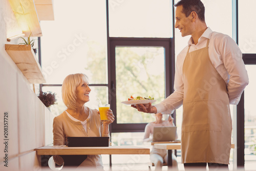 Your order. Charming young waiter bringing salad to his elderly customer and smiling at her while she drinking juice