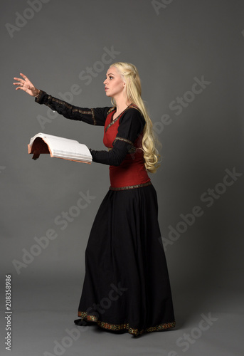  full length portrait of pretty blonde lady wearing a red and black fantasy medieval gown, holding a book. standing pose on grey background.