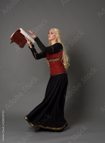  full length portrait of pretty blonde lady wearing a red and black fantasy medieval gown, holding a book. standing pose on grey background.