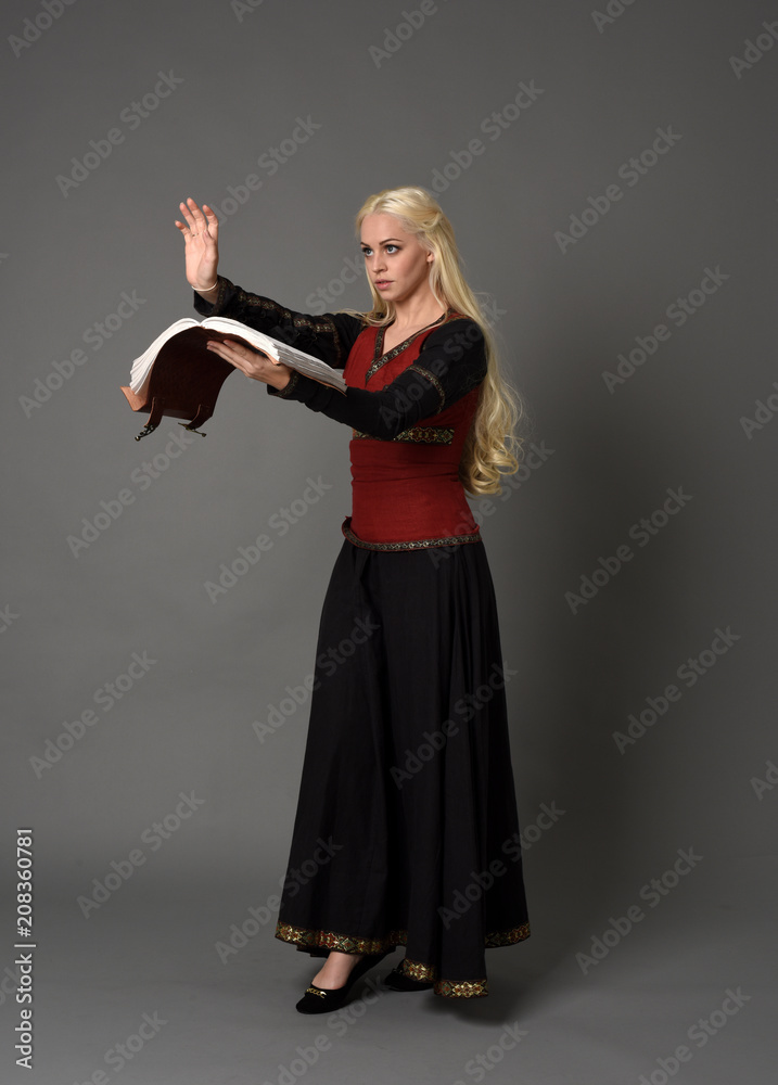  full length portrait of pretty blonde lady wearing  a red and black fantasy medieval gown, holding a book. standing pose on grey background.