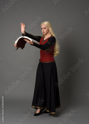  full length portrait of pretty blonde lady wearing a red and black fantasy medieval gown, holding a book. standing pose on grey background.