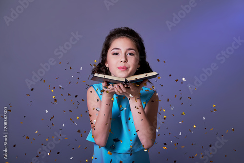 Young woman or teen girl is blowing glitter confetti.Asian girl teenager holds book in hands,on purple background festive sparkles for the party are flying.Concept Christmas,new year,holiday,birthday.