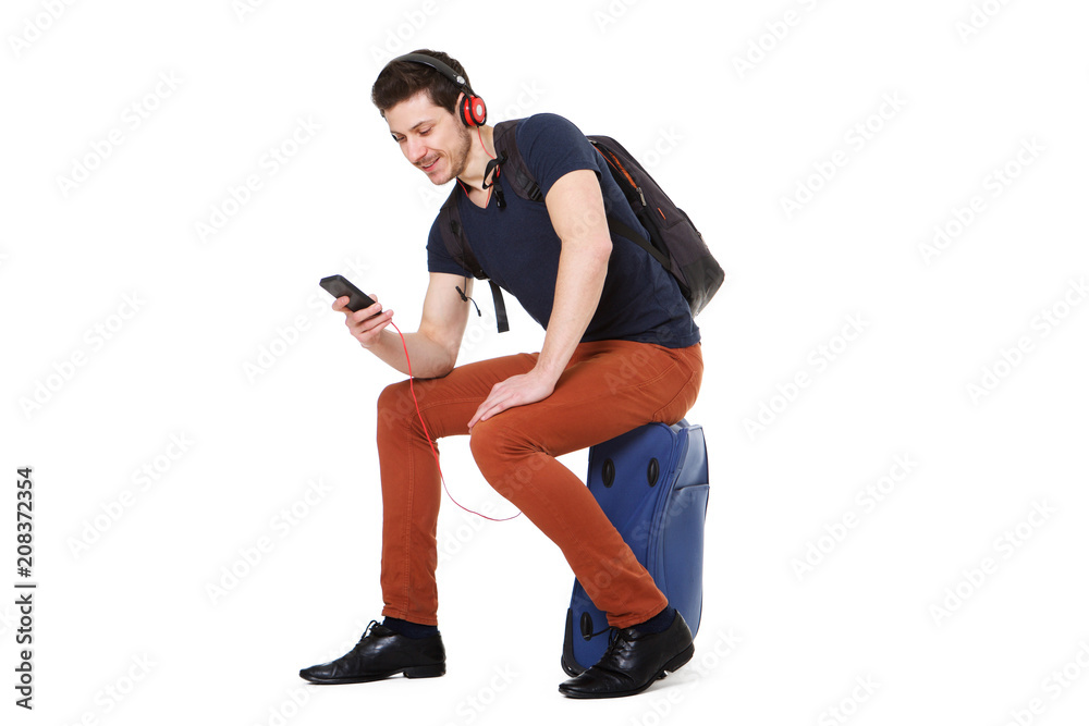 young man sitting on suitcase and looking at mobile phone while listening to music young man sitting on suitcase and looking at mobile phone while listening to music