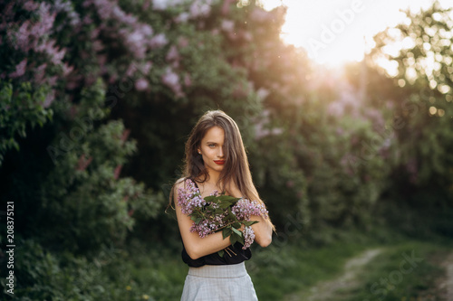 The charming girl stands in the park and keeps a bouquet