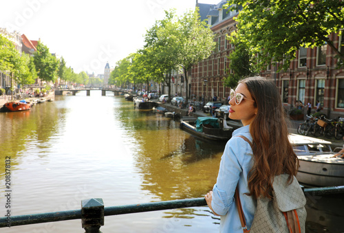 Portrait of traveler girl with sunglasses and backpack enjoying Amsterdam city. Back view of young woman looking to the side on Amsterdam channel, Netherlands, Europe.