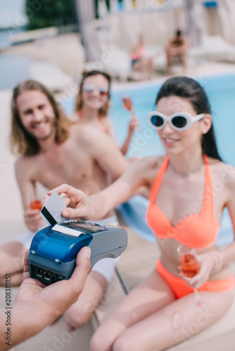 smiling young woman paying with credit card while drinking champagne with friends at poolside