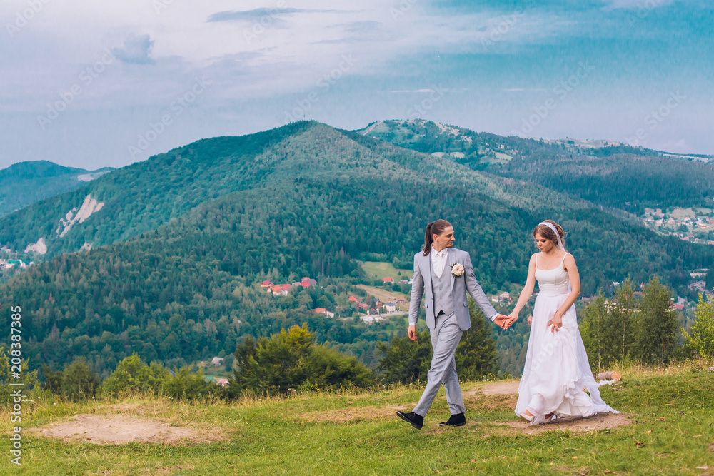 Wedding couple running along the grass against the background of mountains. Bride and groom holding hands and running to the hill. Wedding day of young couple outdoor.
