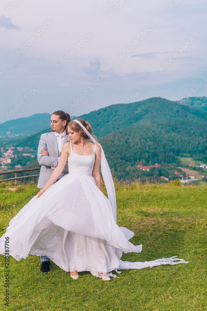 Handsome groom is gently holding his elegant new wife under pine tree. Mountain landscape. Young couple. Wedding day. 