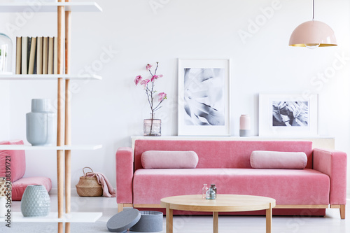 Posters and flowers above pink sofa in pastel living room interior with wooden table. Real photo