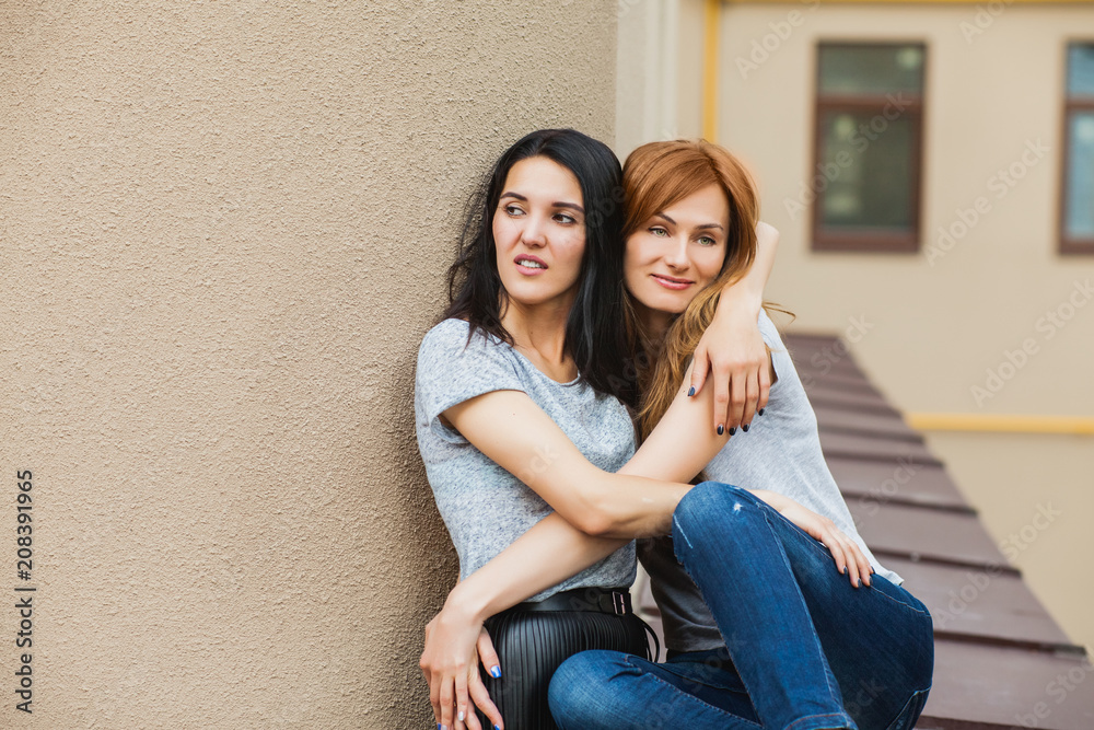 Two cheerful girlfriends hugging near building in the street. one girl with long brunette hair in black skirt, another redhead girl wearing gray shirt and blue jeans. concept of sincere friendship