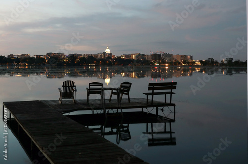 Madison downtown skyline with Wisconsin State Capitol building dome as seen from across lake Monona bay. After sunset sky and downtown skyline reflected in a water. Pier with chairs on a foreground. © Maryna